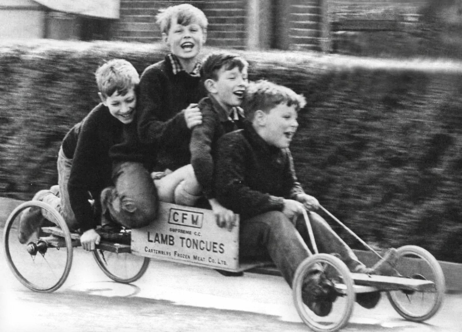 Four boys racing down a street on a home-made cart, archival black-and-white photo