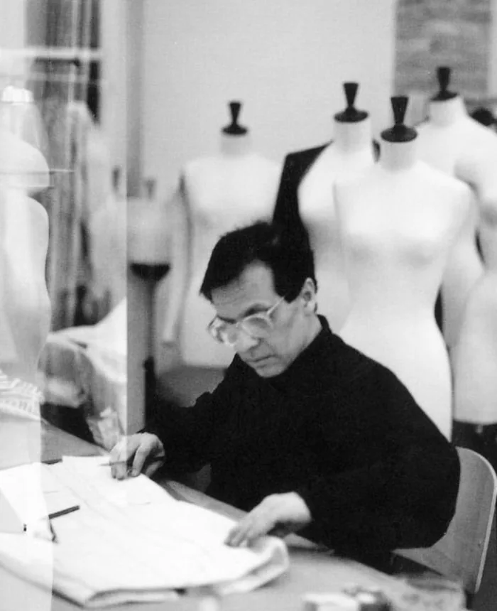 A designer in glasses working on patterns at a table in an atelier, with a row of dress forms behind him