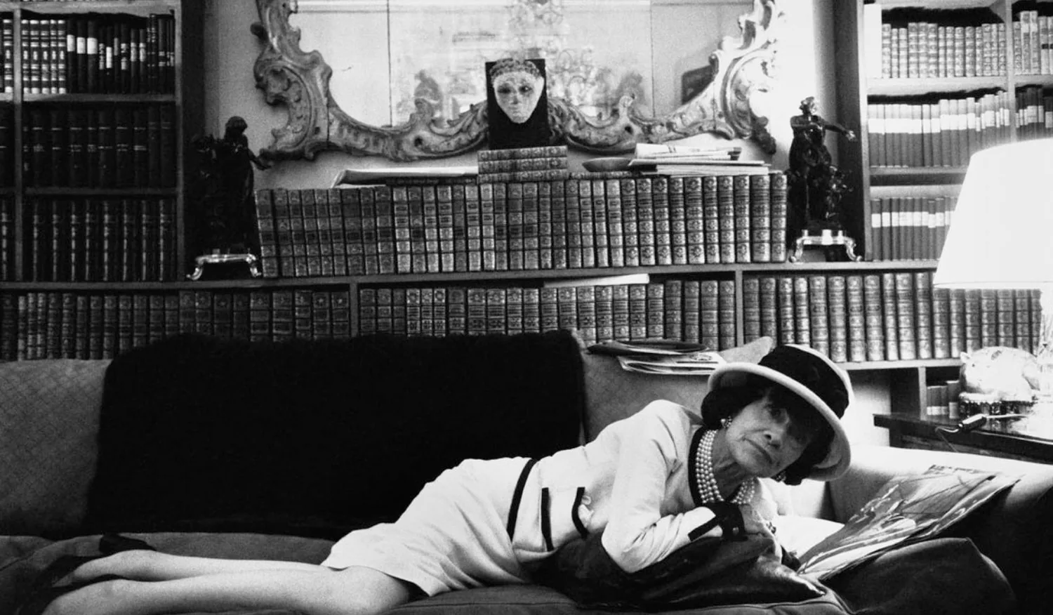 Coco Chanel in a hat and a pale suit, lying on a sofa in front of bookshelves, black and white