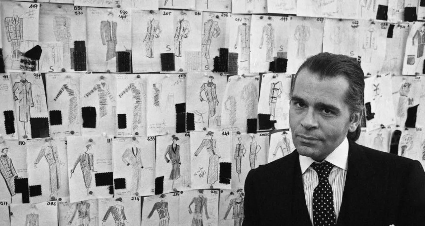Karl Lagerfeld in front of a wall of numbered collection sketches with fabric swatches pinned to them