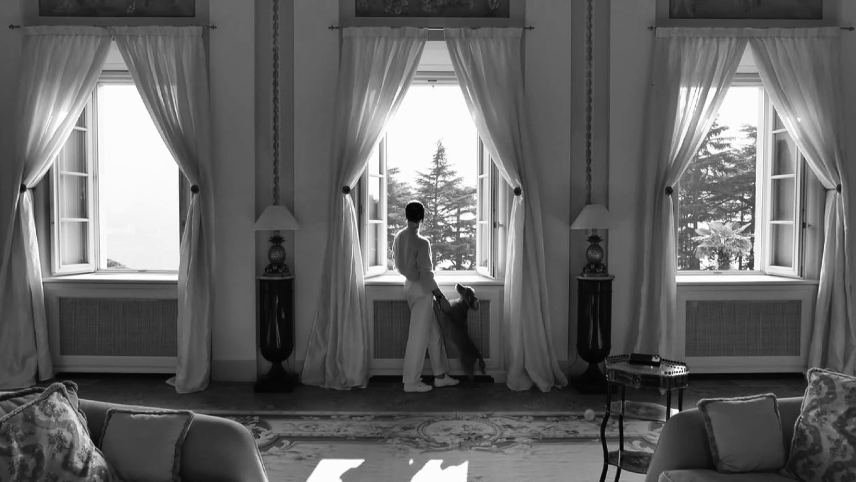 The drawing room of an old villa with three tall windows: a figure with a dog looks out at the garden, black and white
