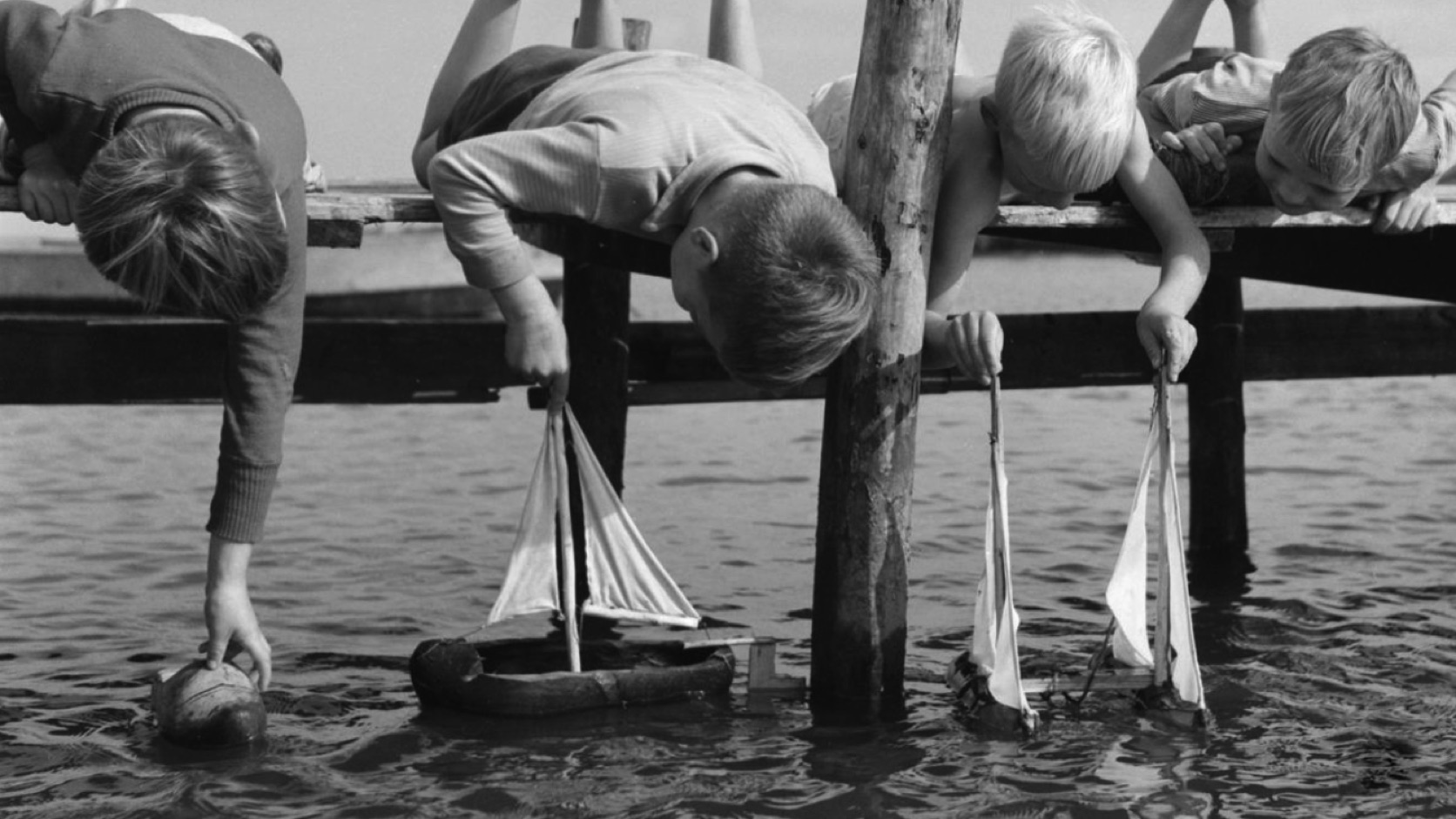 Children lying on a wooden jetty and launching toy sailing boats