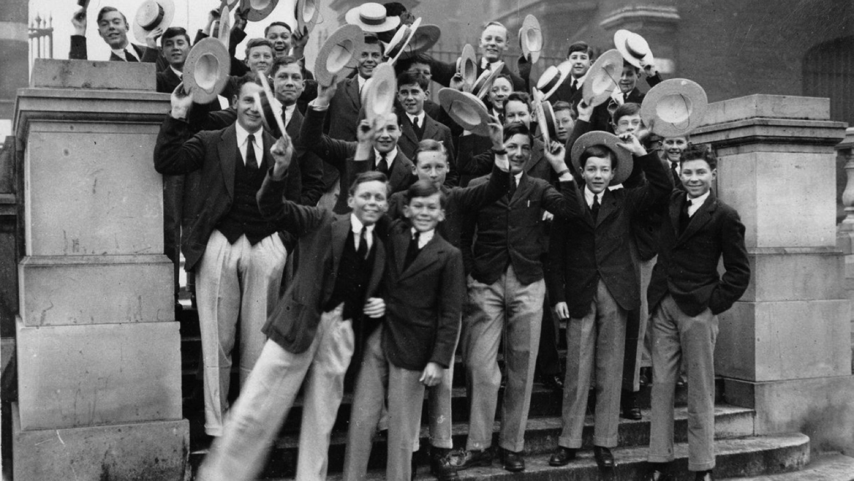 Schoolboys in uniform on the steps of a building raising their straw hats